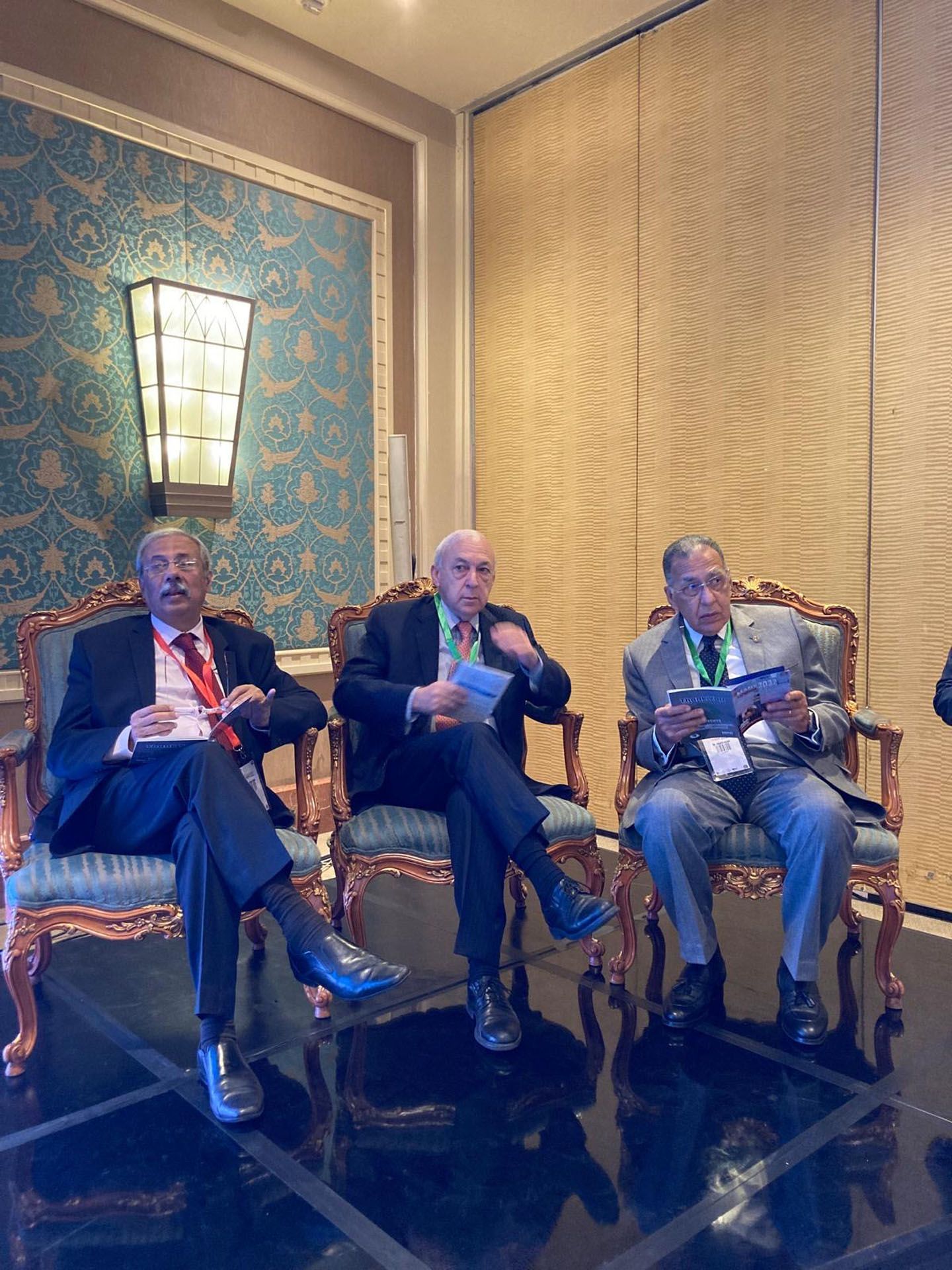 Three men seated, holding papers, in ornate chairs. Conference setting, gold and teal decor.