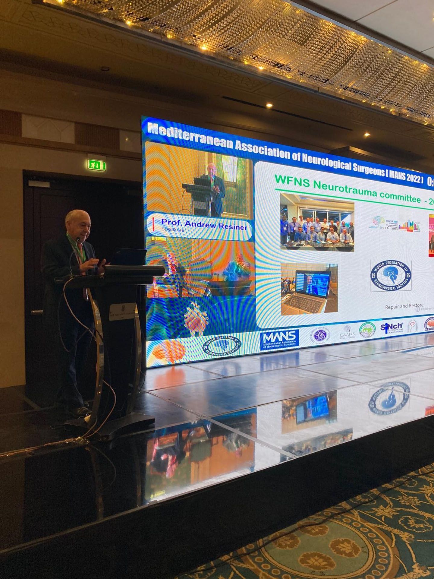 Man presenting at conference with large screen displaying information and logos.