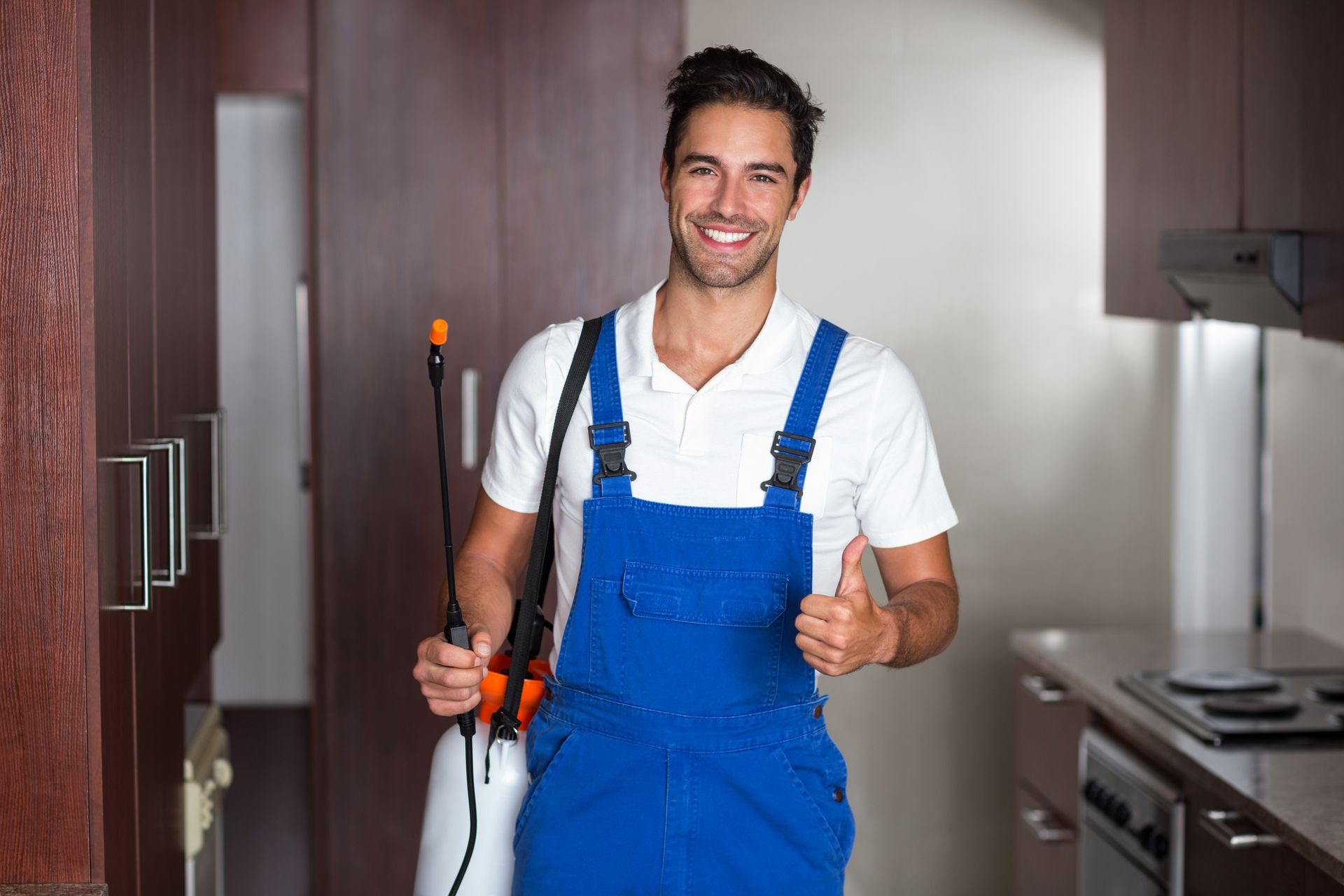 A pest control technician in blue coveralls holding a spray wand gives a thumbs up in a kitchen.