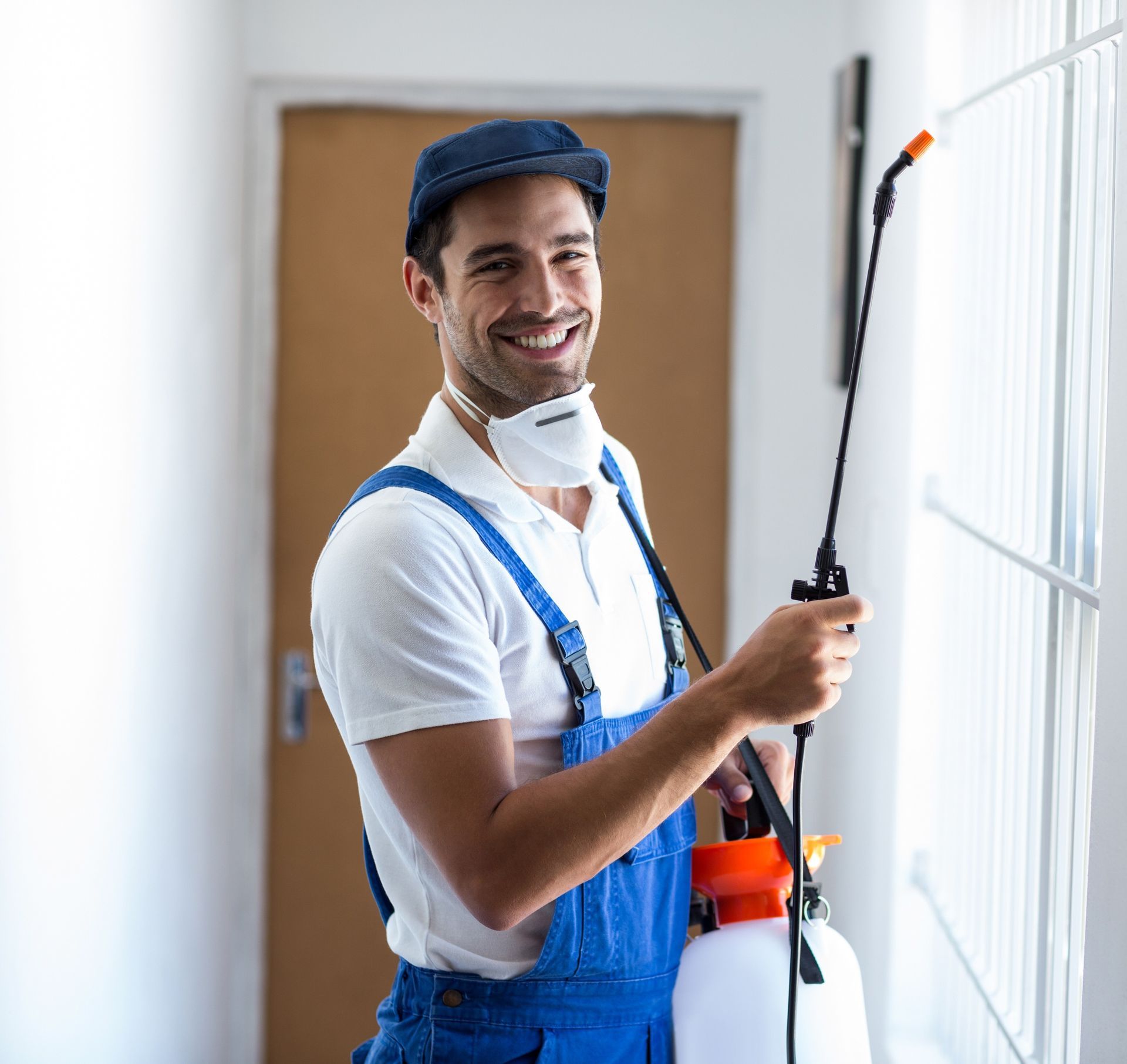 A smiling pest control worker in a blue uniform and face mask sprays a chemical solution near a window.
