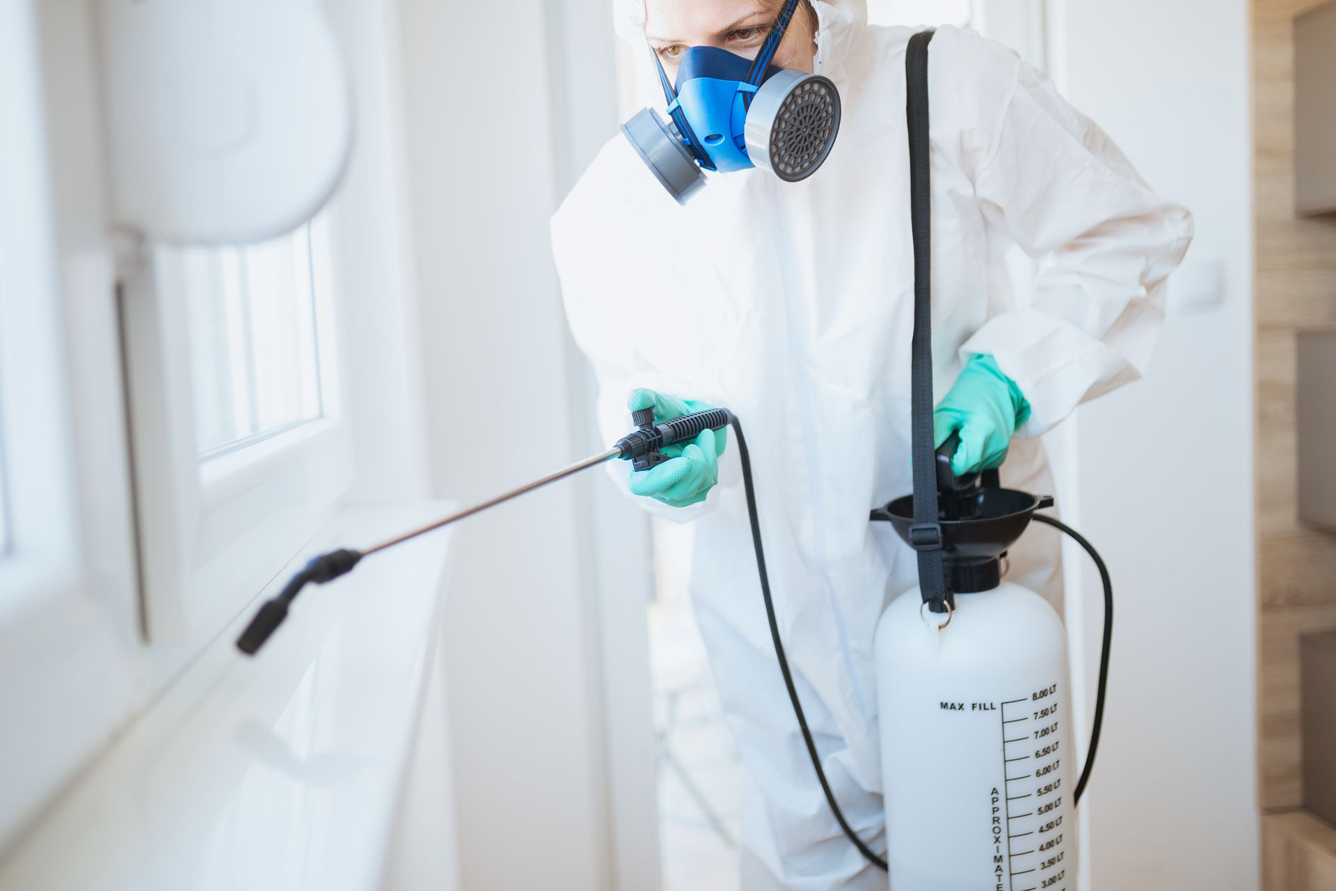 A person in protective gear and a respirator uses a handheld sprayer to treat a window frame in a room.