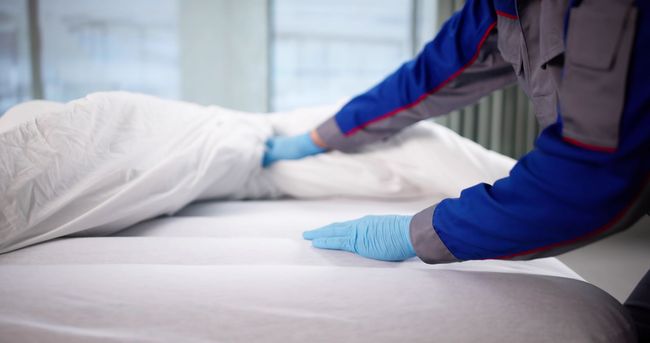 A person in a blue and grey uniform with blue gloves makes a white bed in a bright, clinical-looking room.