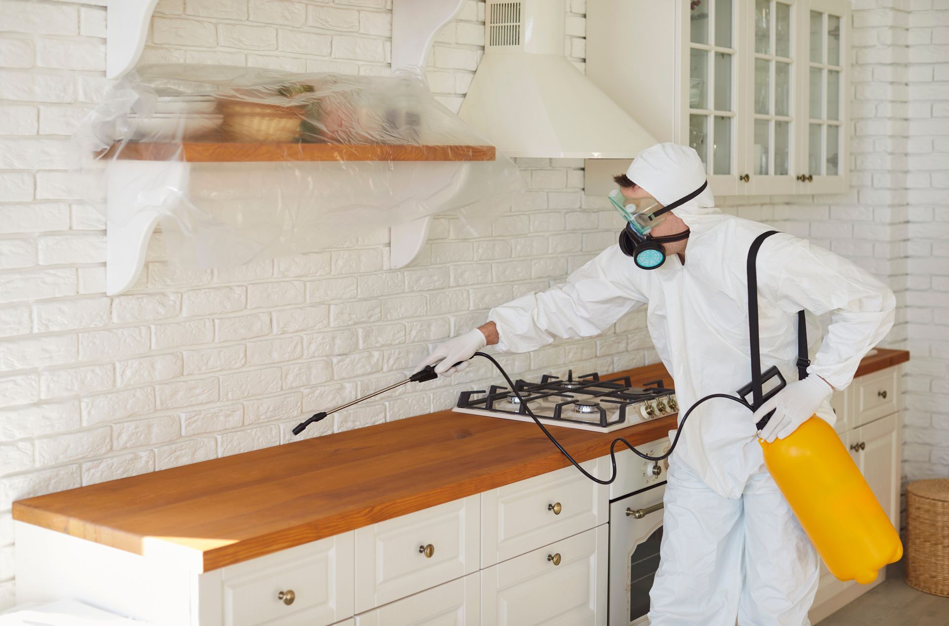 A professional in a white hazmat suit and respirator sprays a kitchen countertop with a yellow chemical tank.