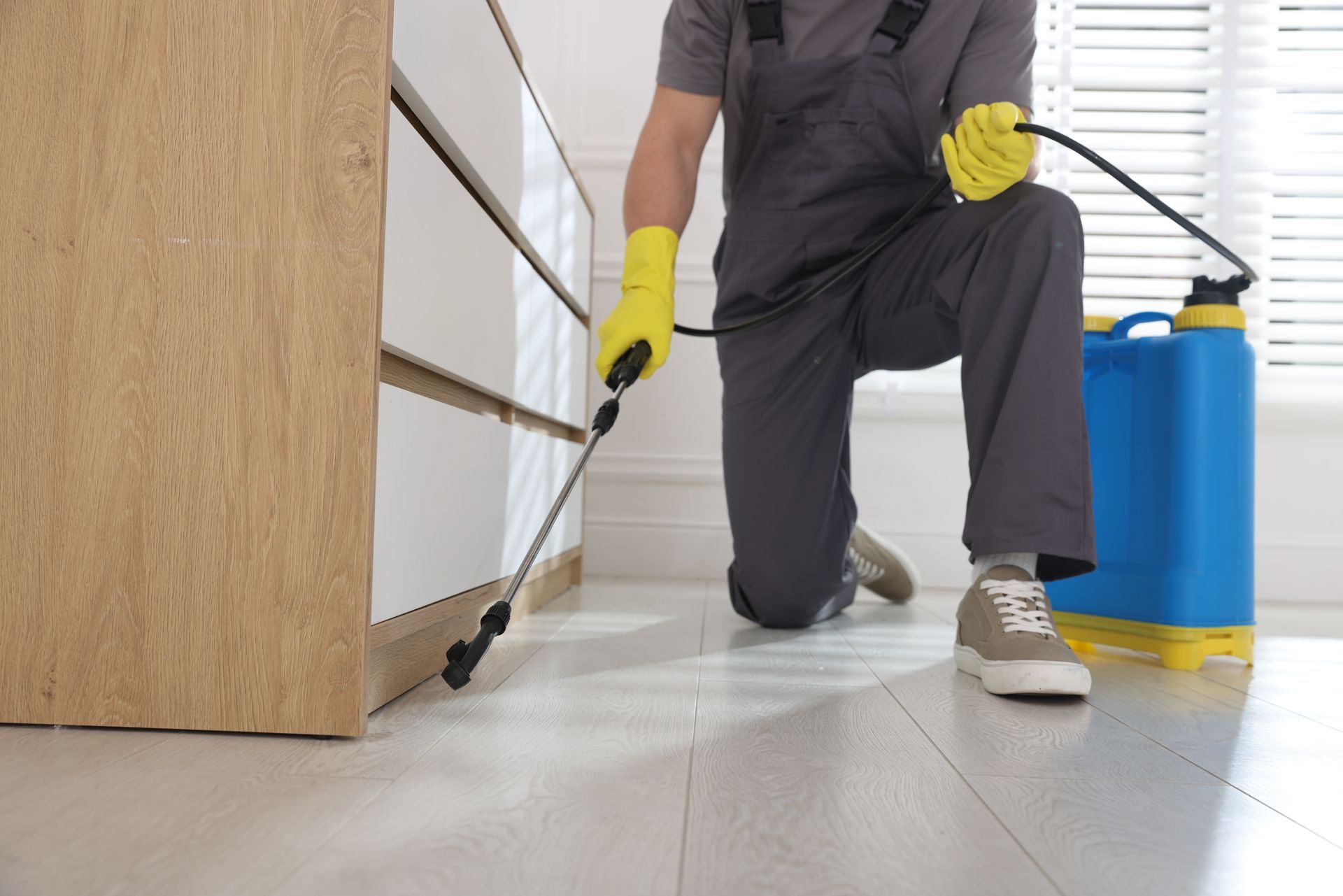 Pest control worker kneeling, spraying insecticide near a cabinet in a room.