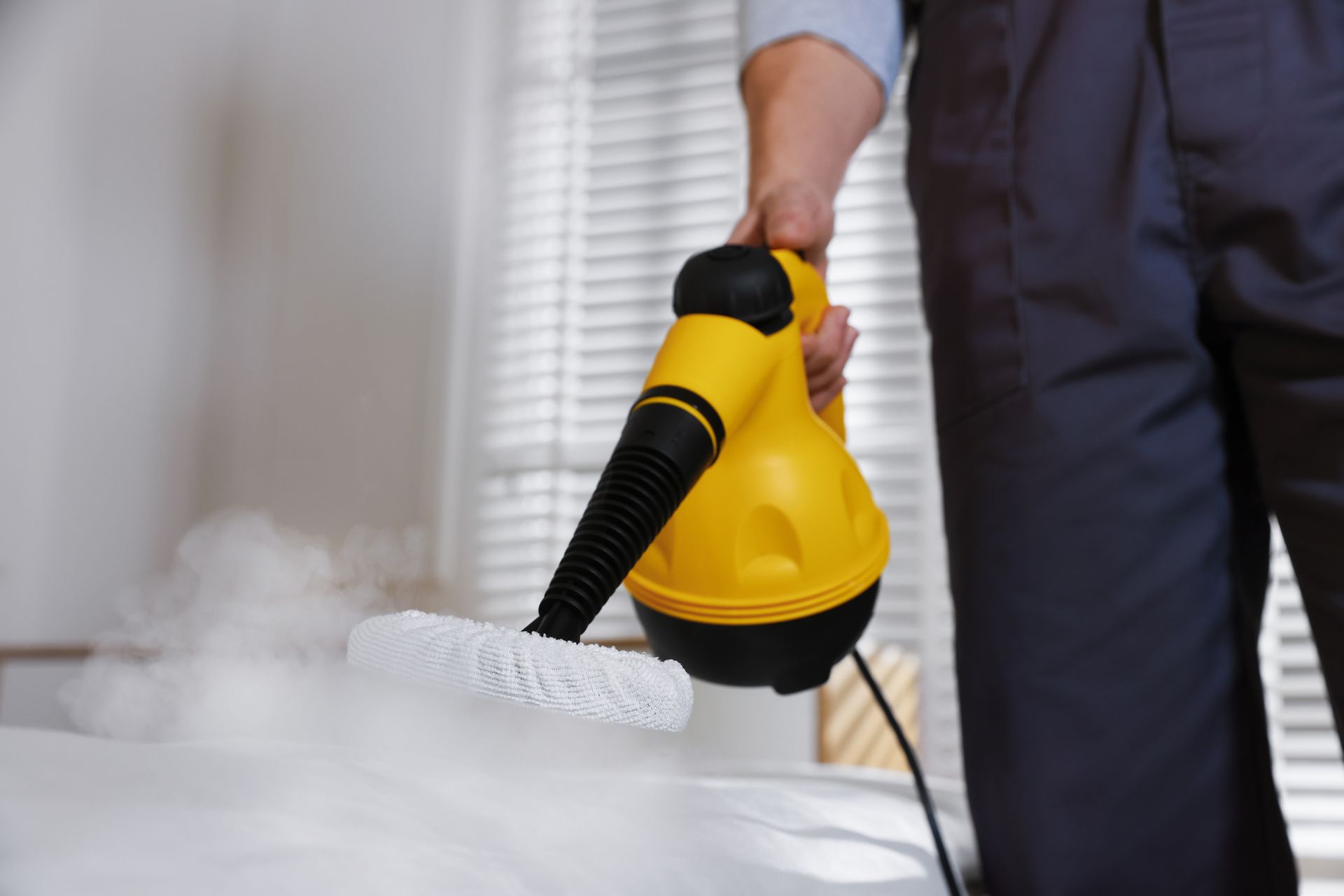 A person in work clothes uses a handheld yellow steam cleaner on a white surface, emitting a cloud of steam.