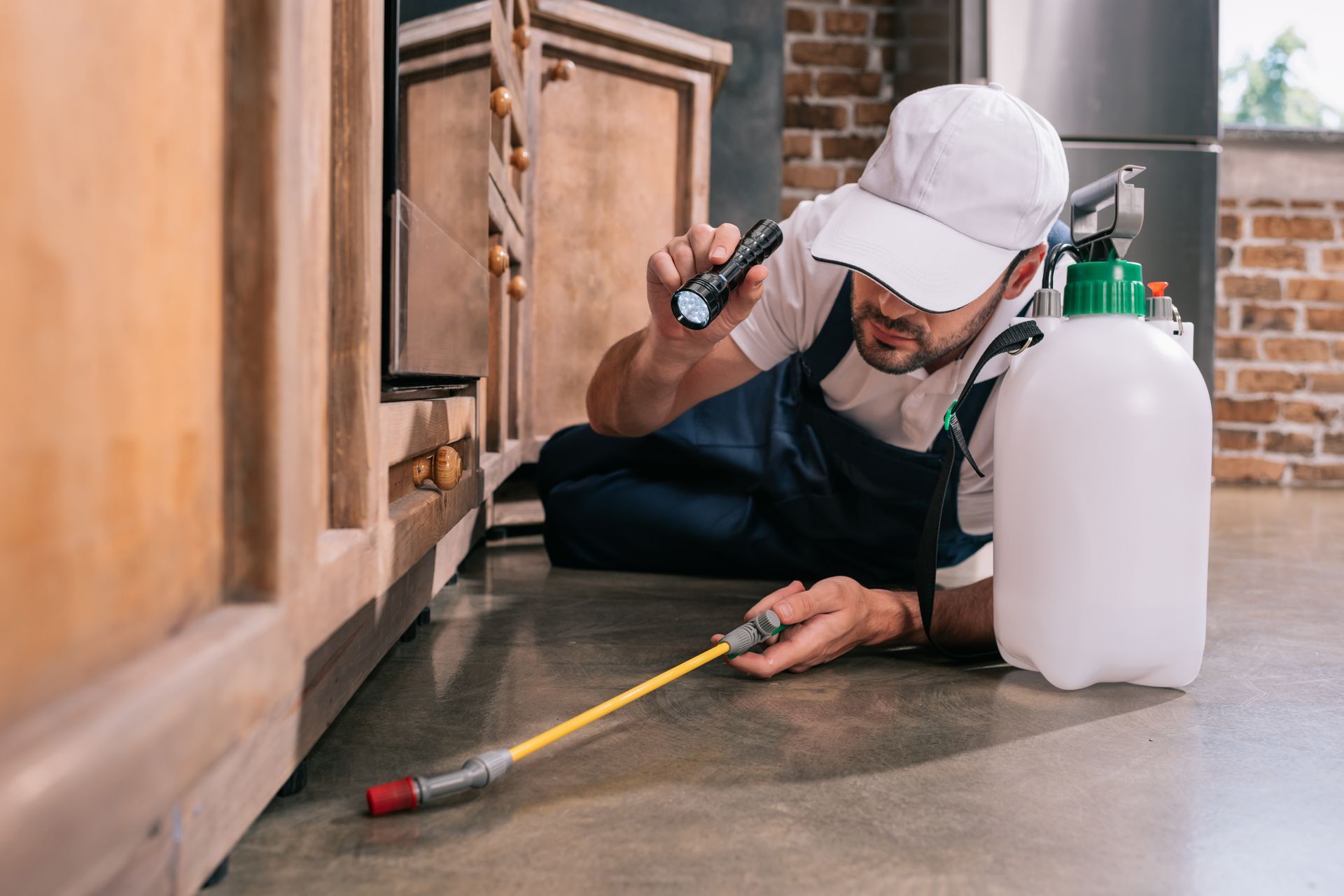 Pest control worker in uniform using a flashlight and spray wand to treat the area under kitchen cabinets.