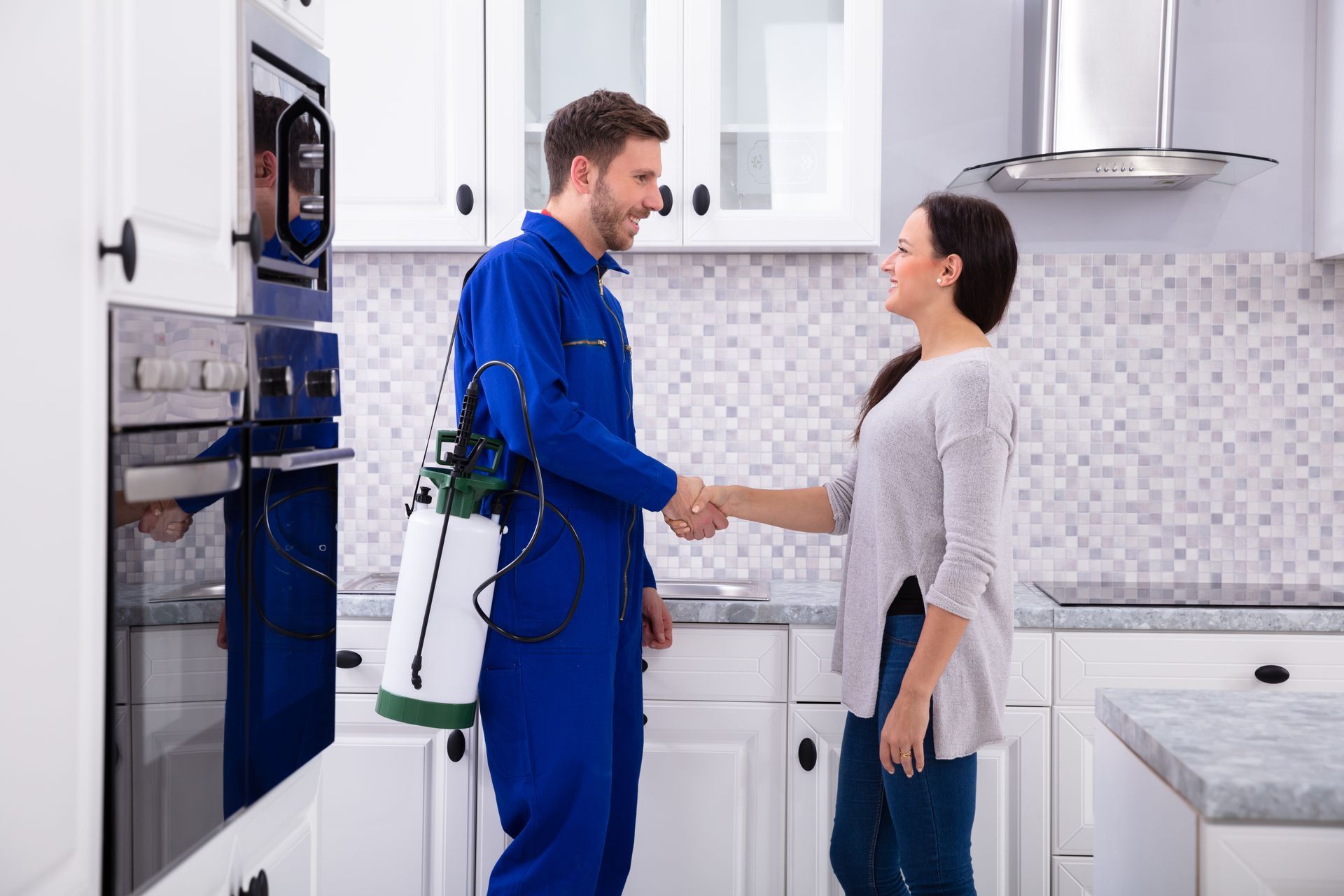 A pest control technician in a blue uniform shakes hands with a homeowner in a bright, modern kitchen.