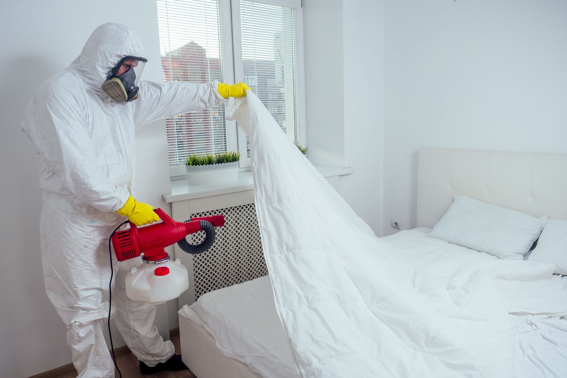 A person in a protective suit and respirator uses a red handheld fogger to treat a bed for pests.