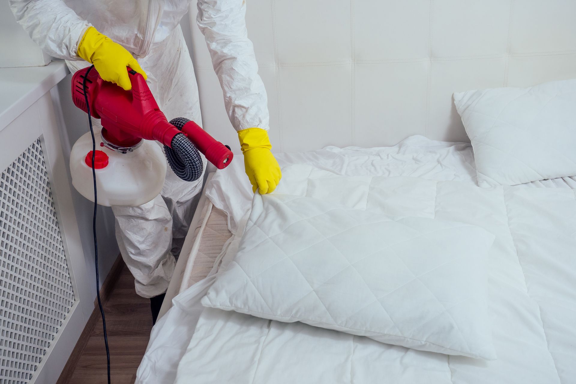 A person in a white hazmat suit and yellow gloves uses a red handheld sprayer to disinfect a white bed.