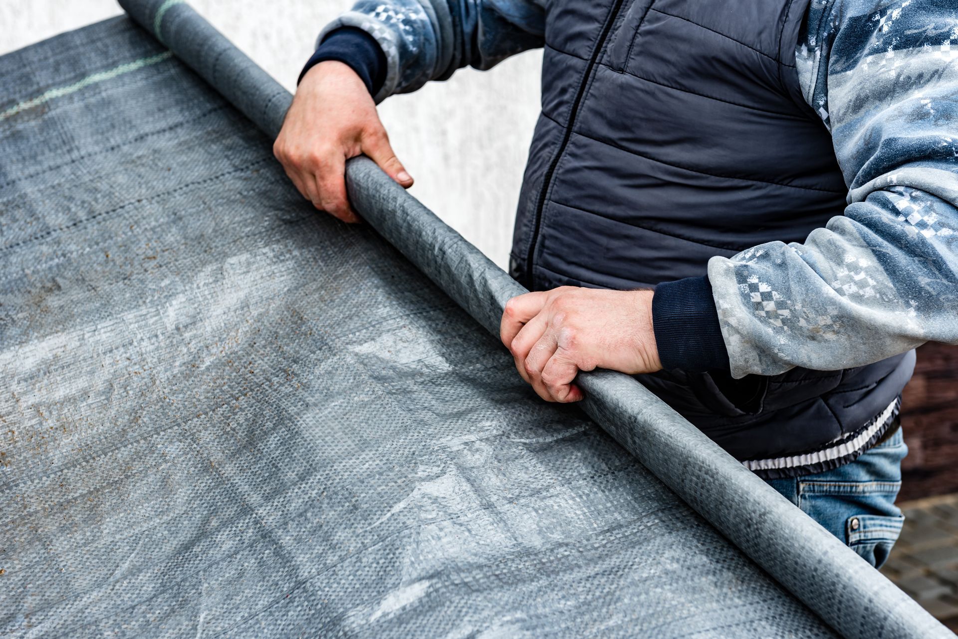 A person in a dark vest unrolls a textured, gray plastic barrier outdoors.