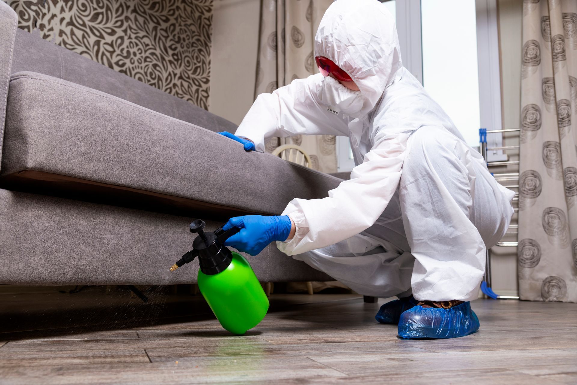 A person in a white hazmat suit and blue gloves disinfects the base of a gray sofa with a green spray bottle.