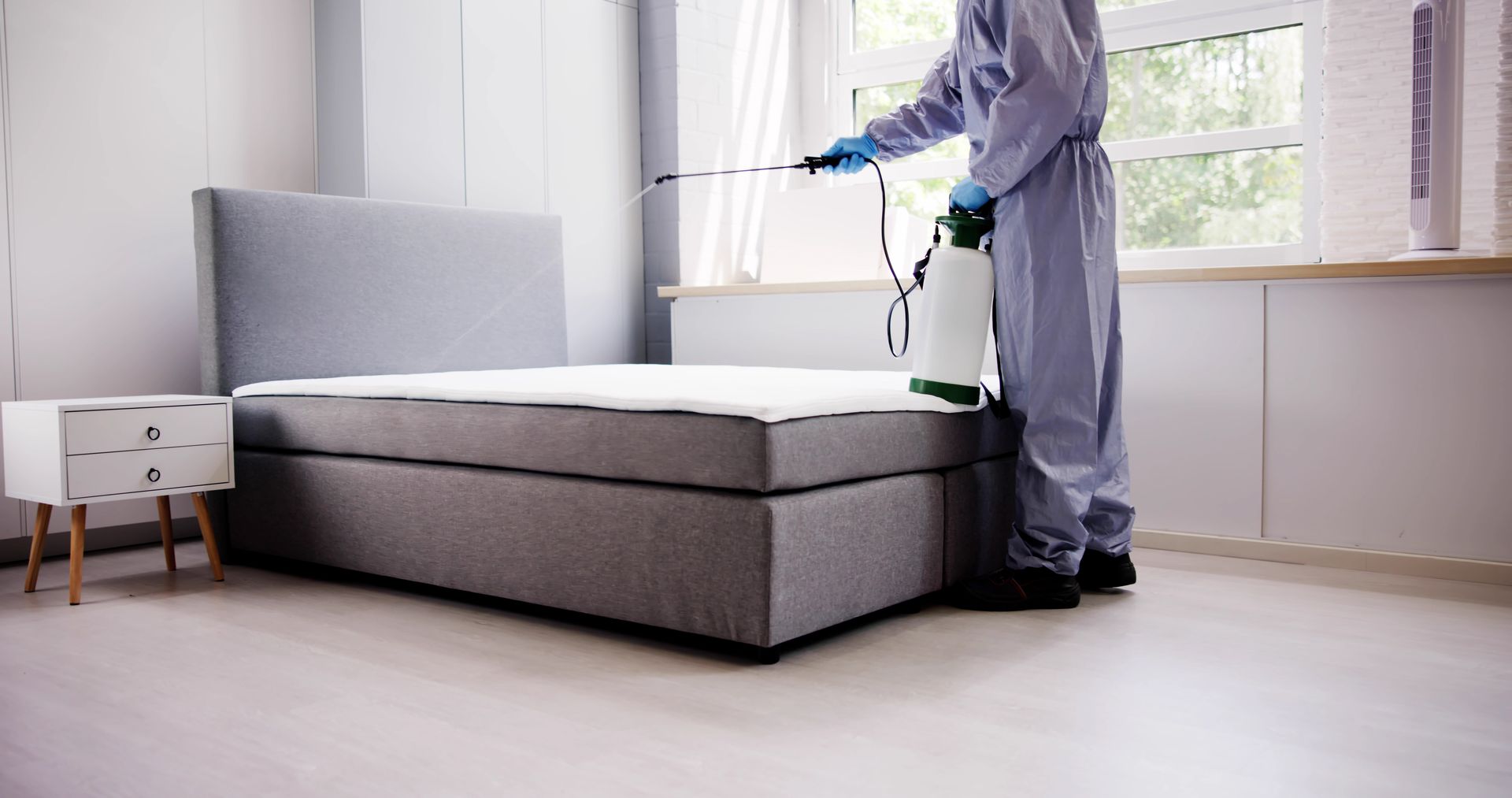 A professional in protective gear uses a handheld sprayer to treat a mattress in a bedroom.