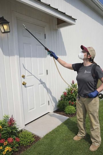 Person spraying insecticide near a white building door; wearing a respirator and safety gear.