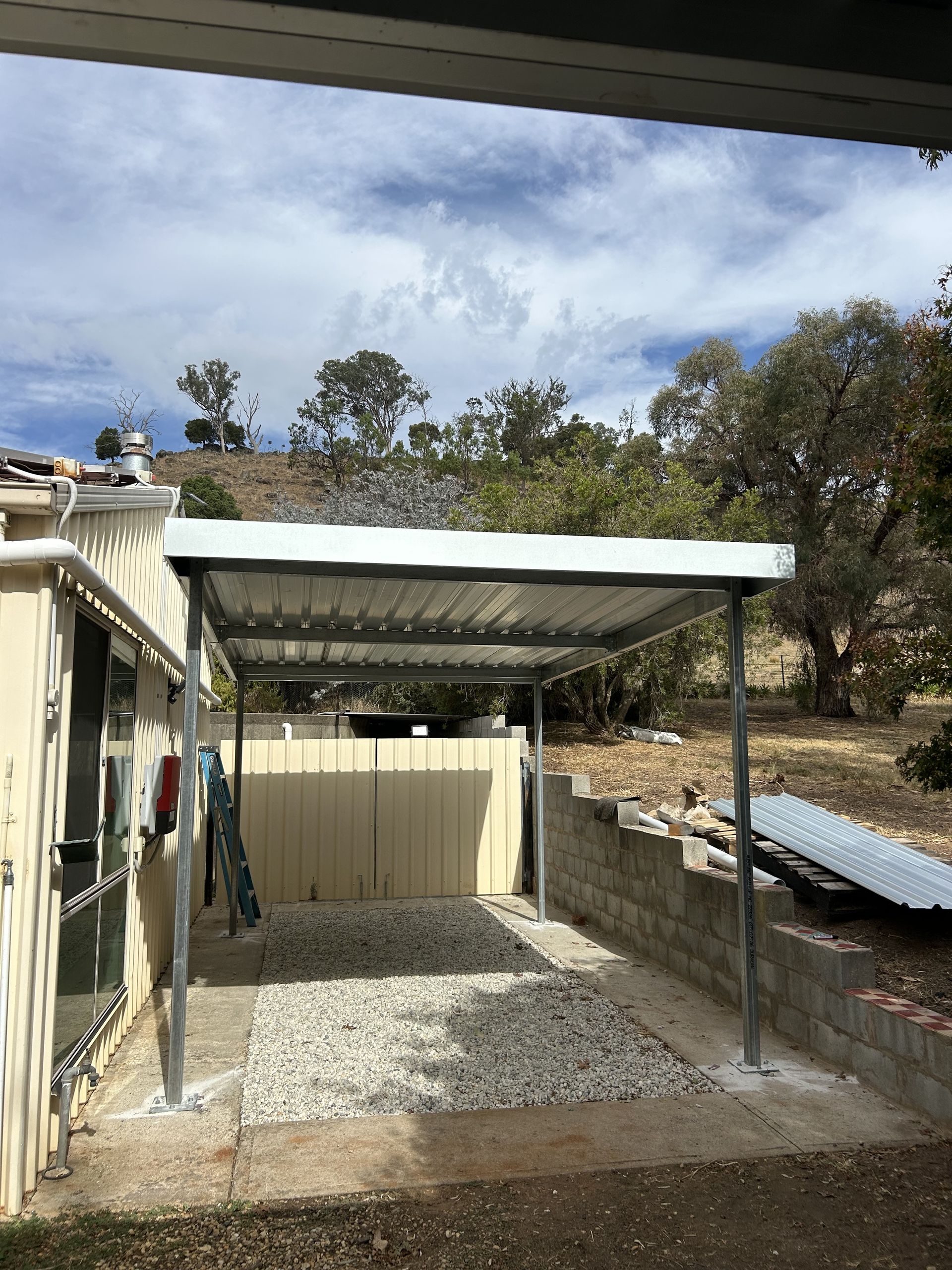 A metal carport structure on gravel, next to a house, with a tree overhead and a hillside in the background — The Wright Way Building In North Albury, NSW