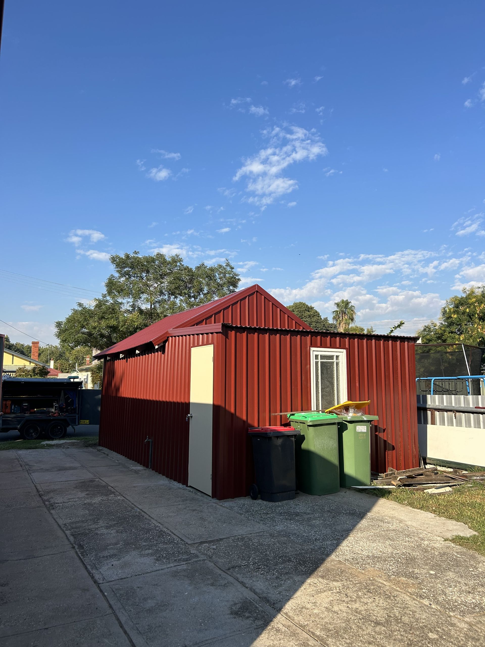 Red metal shed with red roof, two green bins, and a cream-coloured door against a blue sky — The Wright Way Building In North Albury, NSW