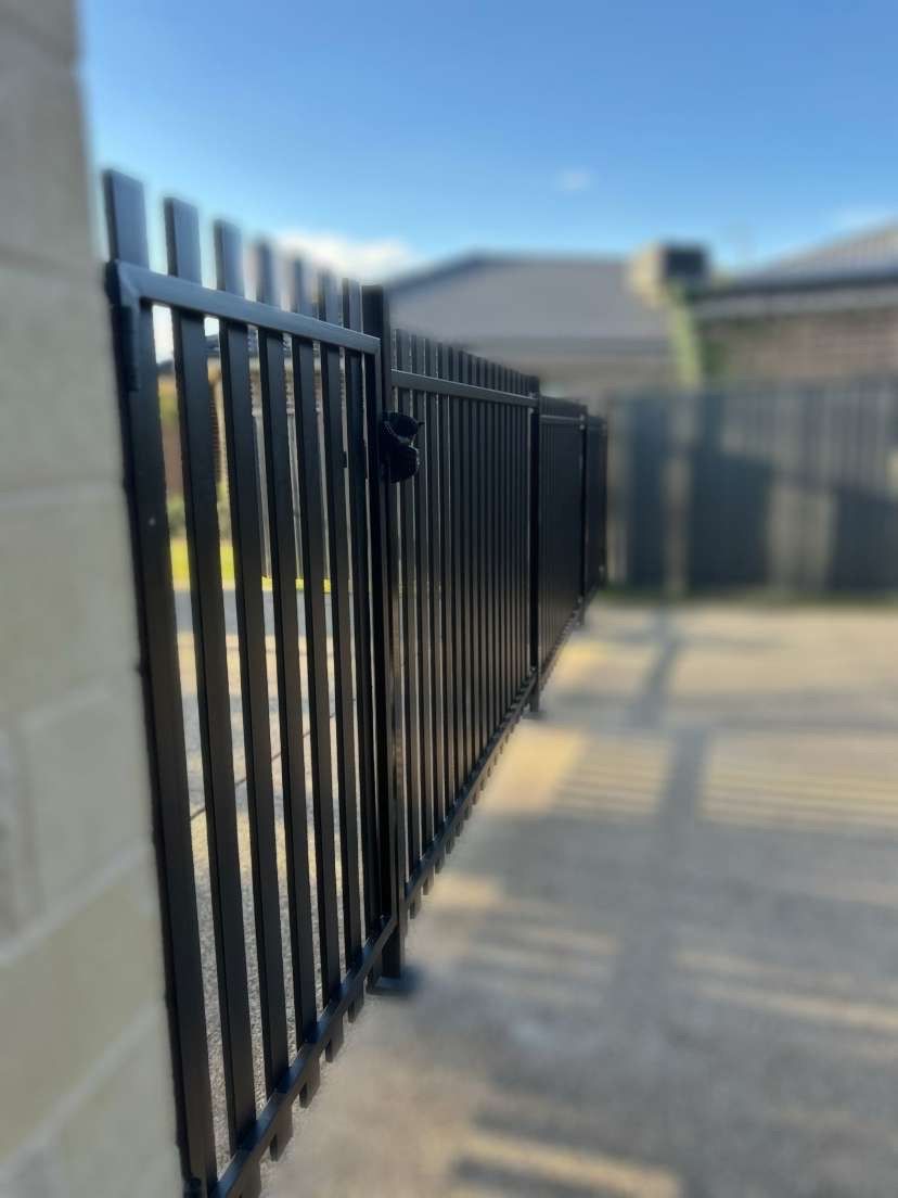 Black metal fence along a beige brick wall, leading into a yard with blurred buildings — The Wright Way Building In North Albury, NSW