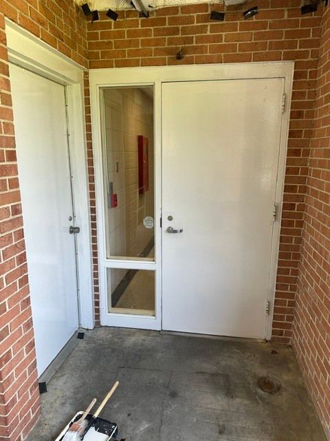 Brick entryway with white doors and a narrow windowed side panel; concrete floor and brick walls.