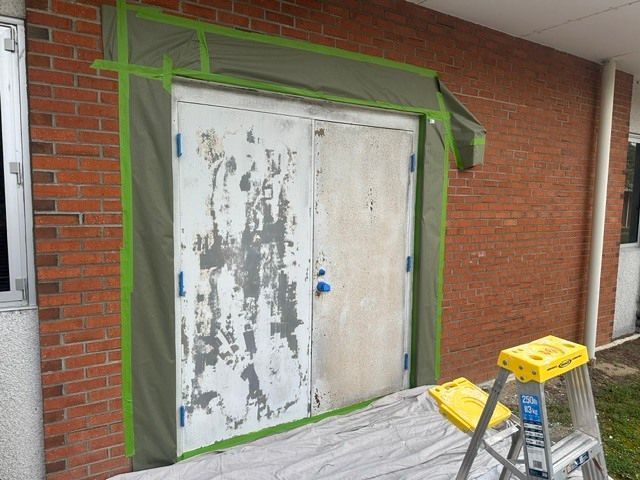 Boarded-up double doors on a brick wall, masked with green painter’s tape and surrounded by construction materials.