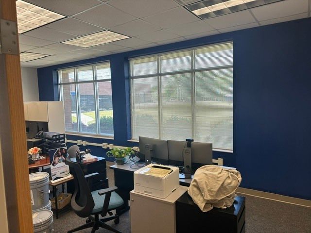 Office workspace with blue walls, desks, computers, chairs, and large windows with blinds