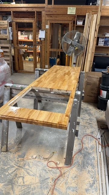 Workshop table frame with a wooden top and a fan in the background