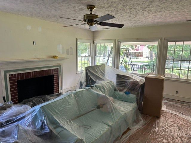 Living room covered in plastic sheeting, with fireplace, ceiling fan, and large windows.