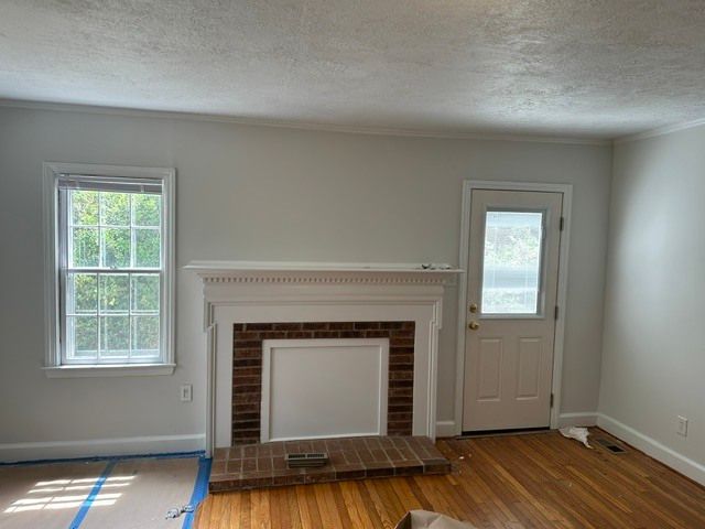 Empty living room with brick fireplace, white walls, wood floor, and two windows and a door.