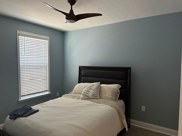 Neatly made bedroom with blue walls, dark bed frame, window blinds, and ceiling fan.