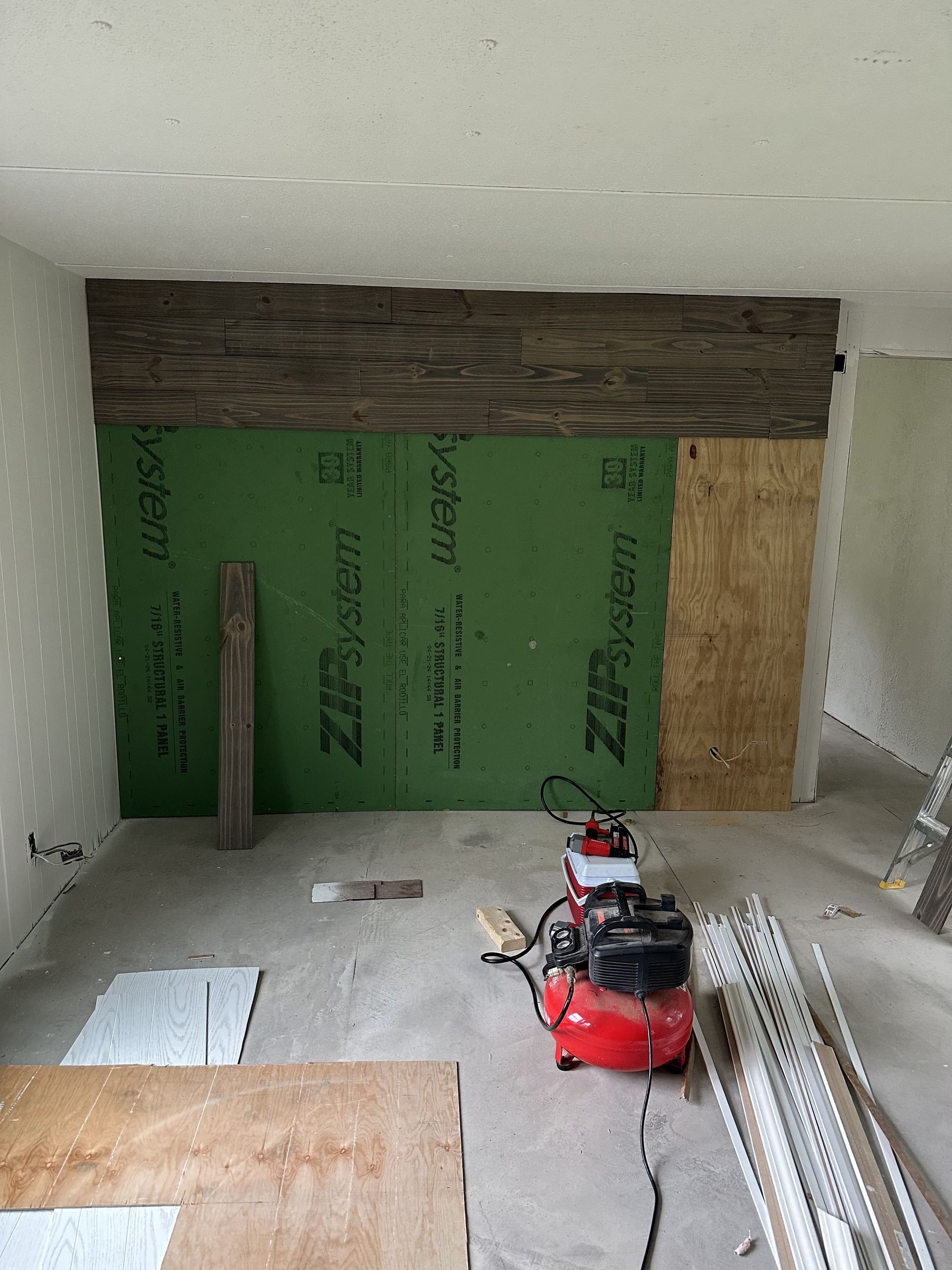 Unfinished room under renovation with green wall insulation, exposed wood, and a red air compressor on the floor.