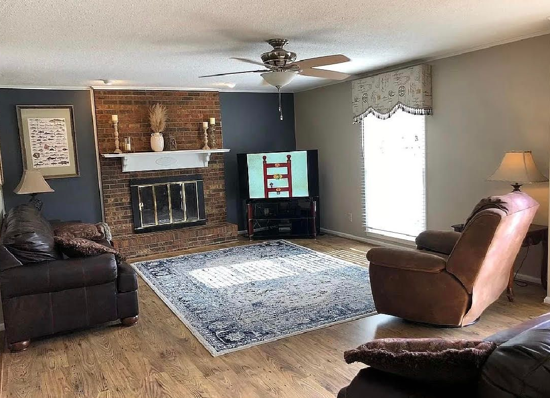 Living room with brick fireplace, TV, brown seating, hardwood floor, and bright window blinds