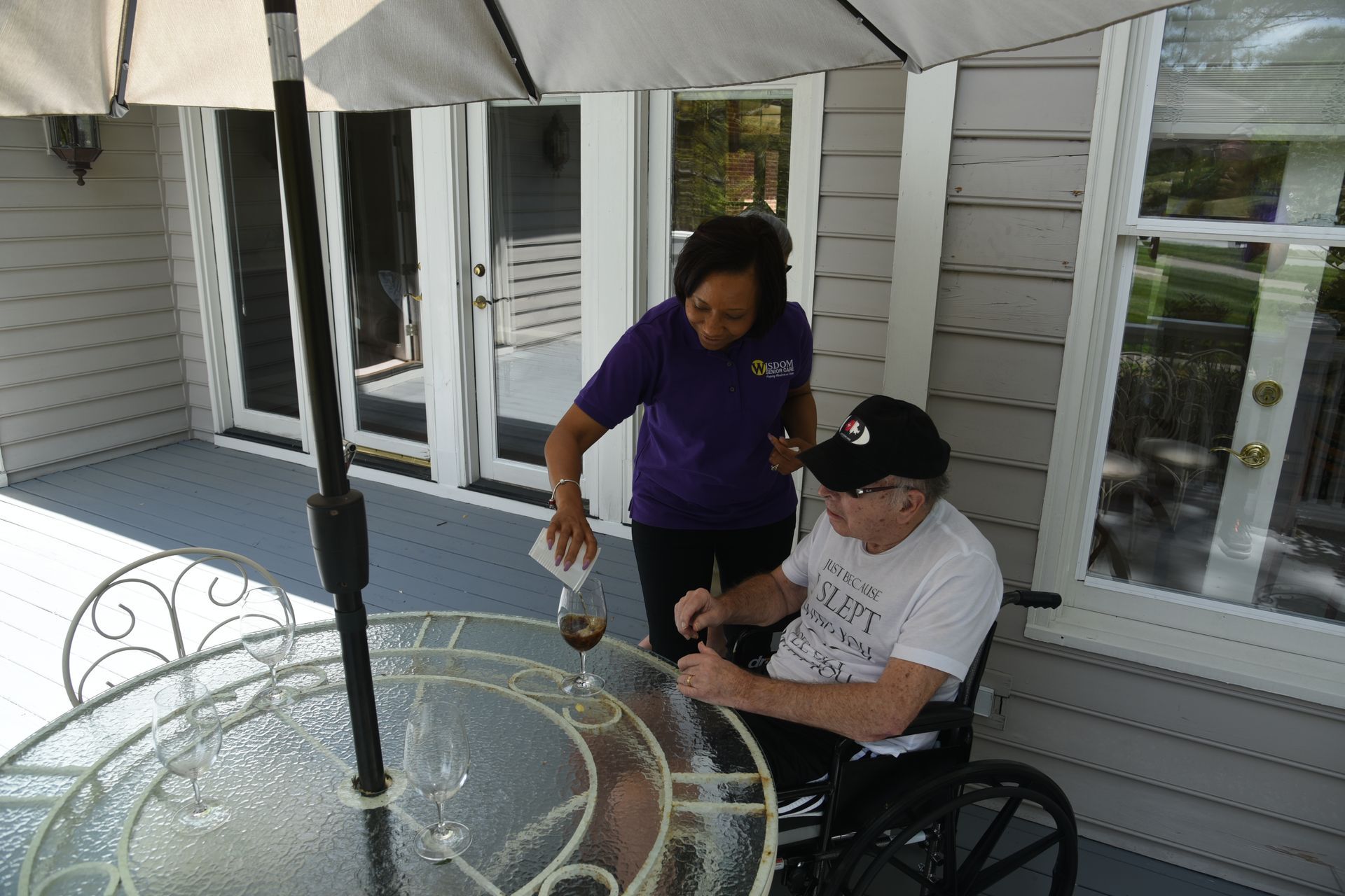 Woman in purple shirt pours a drink for a person in a wheelchair on a patio, under an umbrella.