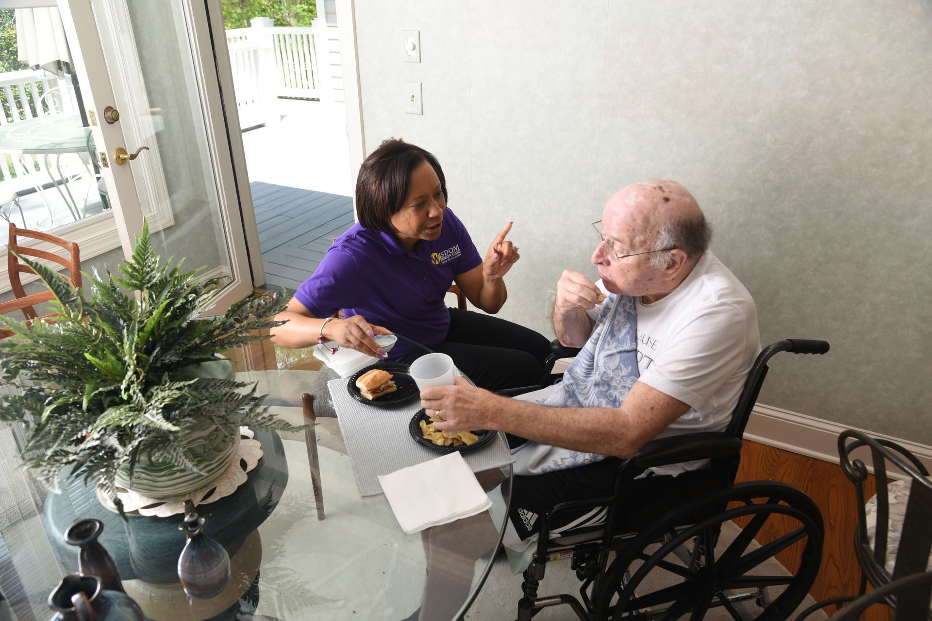 Caregiver assists person in wheelchair with meal on patio.