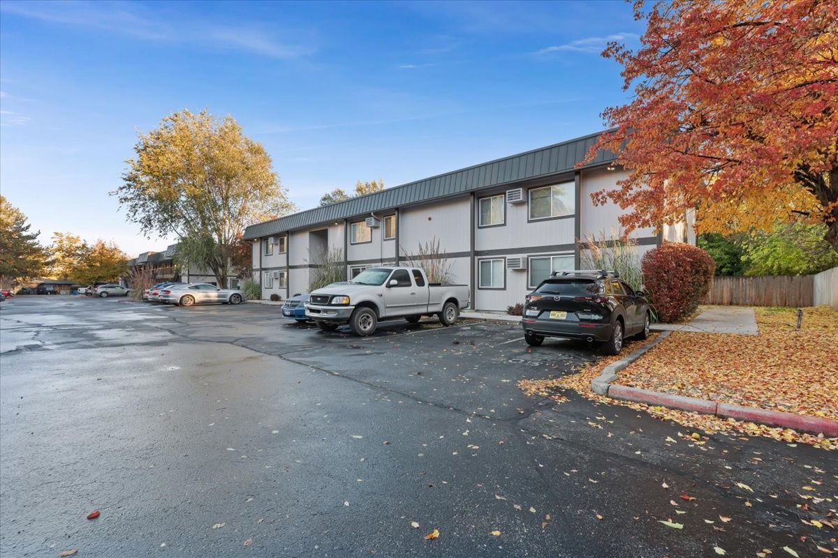 Apartment building with parked cars on an asphalt lot; fall foliage in the background under a blue sky.