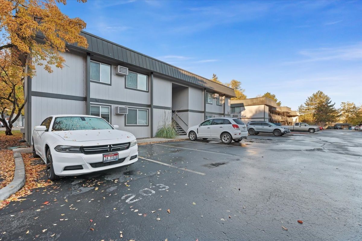 Two-story apartment building with parked cars on a wet, paved lot under a blue sky.