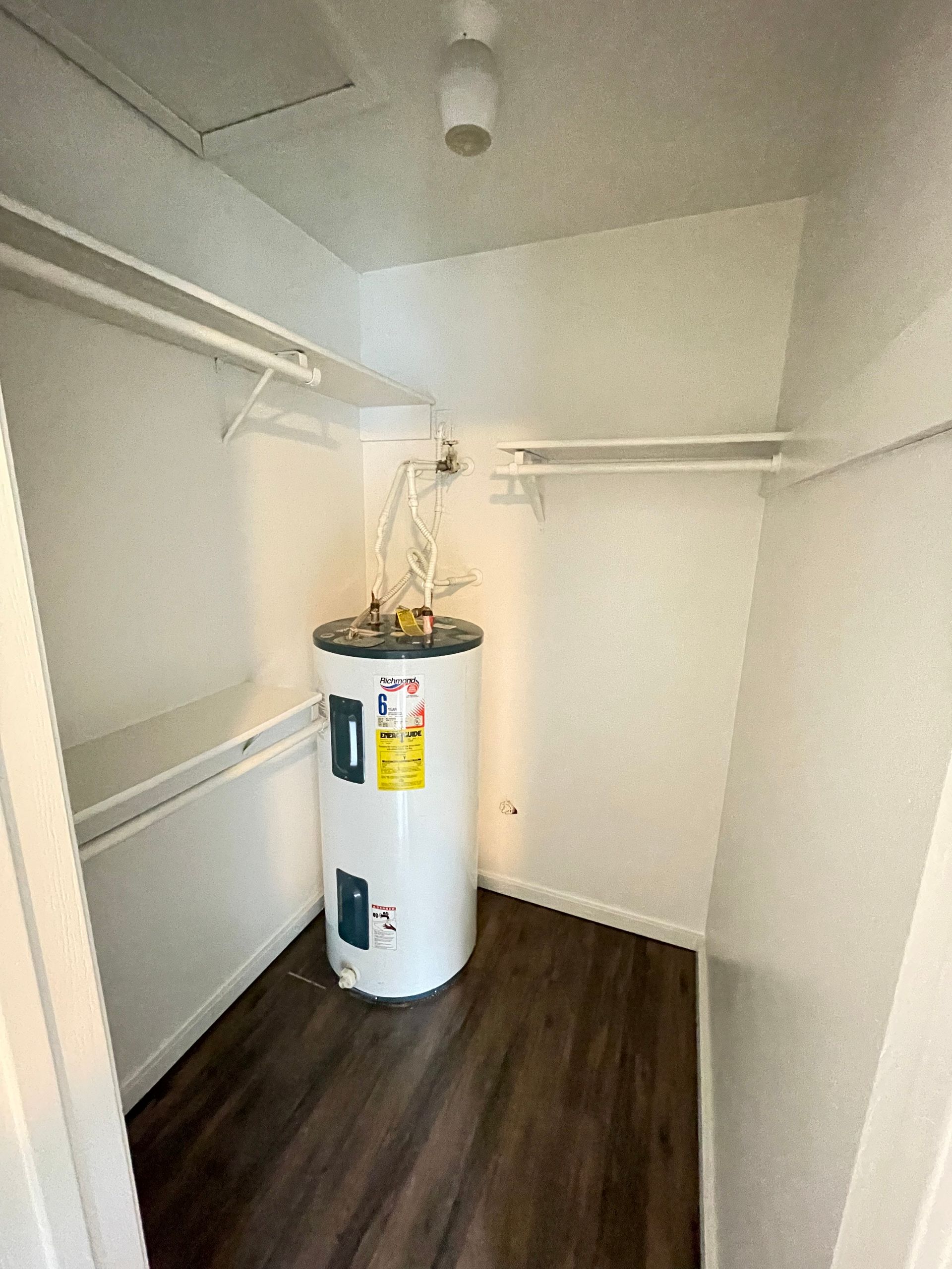A small closet with a water heater on a dark wood floor. White walls and clothes rods.
