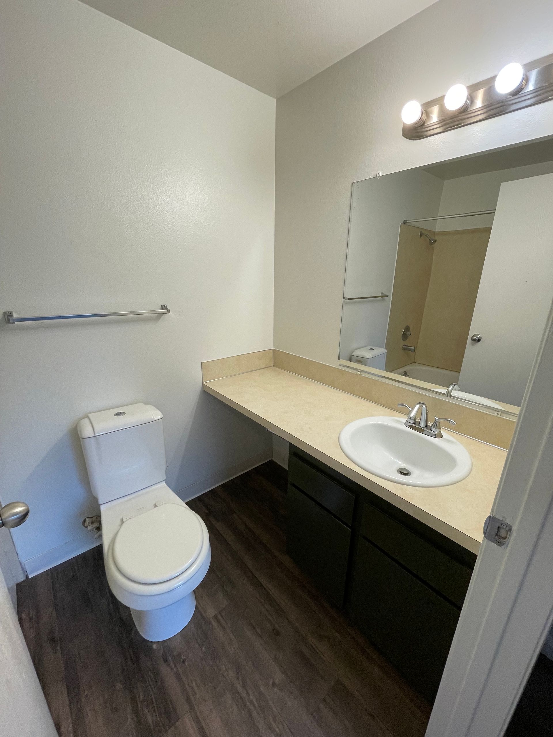 Bathroom with toilet, sink, mirror, and towel rack. Dark wood floor and light countertop.