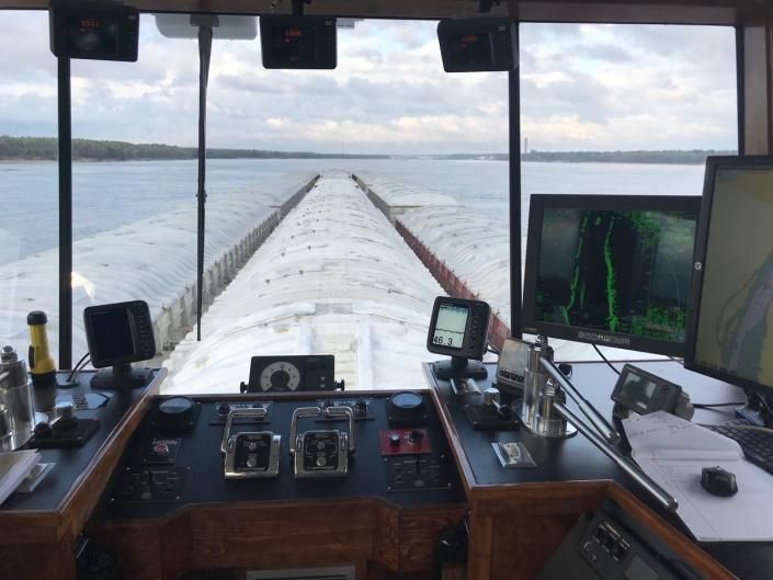 View from a ship's bridge, looking down a long line of barges on water, with navigation instruments.