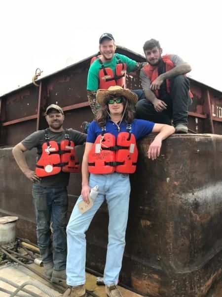 Four people wearing orange life vests pose on a barge.