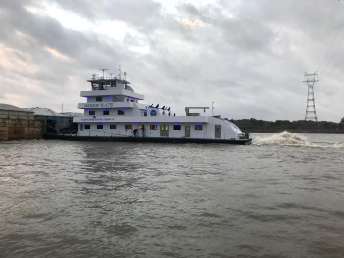 White tugboat on river under cloudy sky, churning water, power line visible.