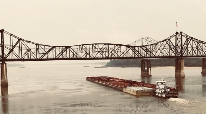 Barge pushing cargo under a steel truss bridge over a river. Cloudy sky.