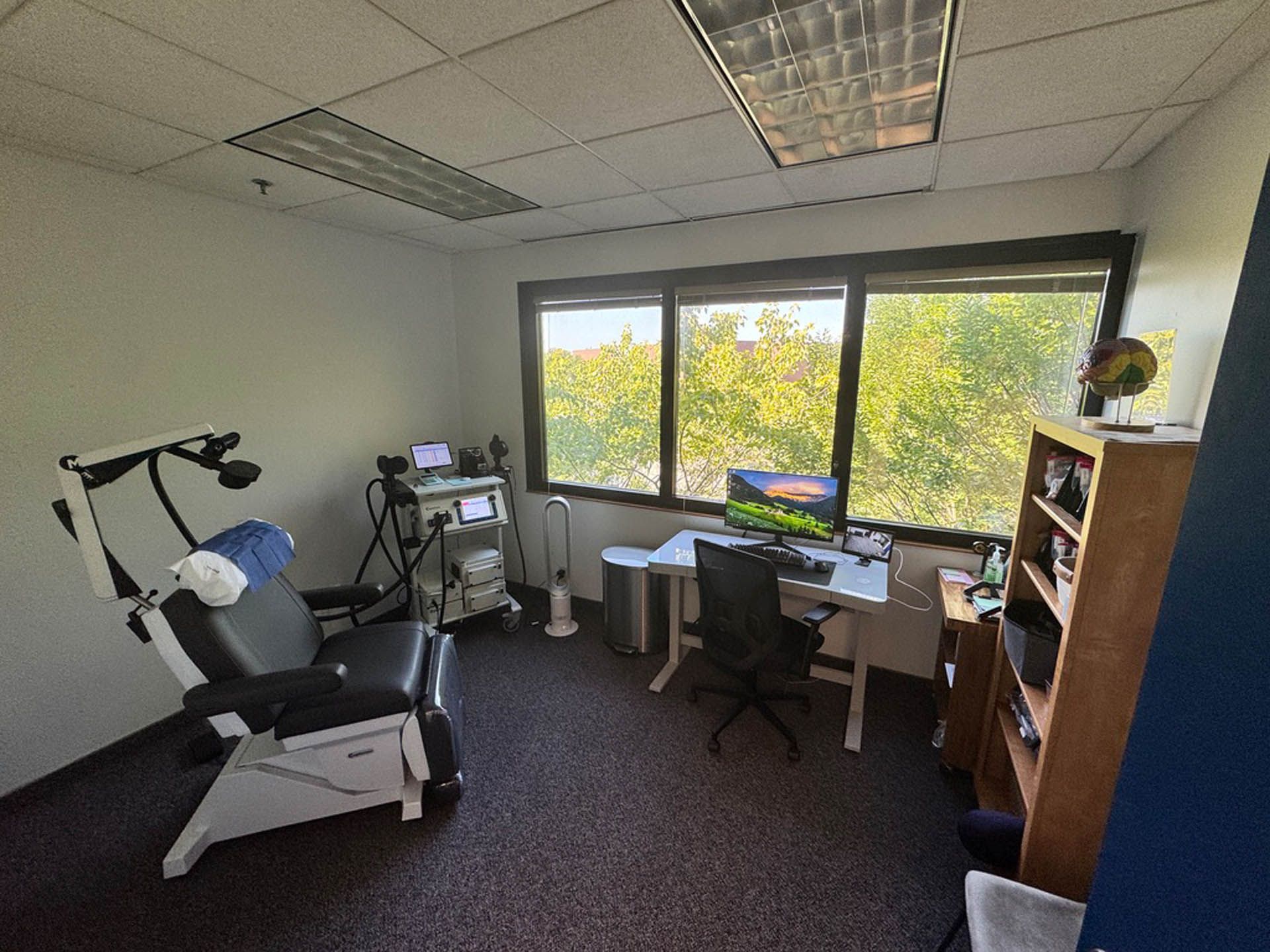 Medical office: a reclined chair with equipment, desk with computer near a window overlooking trees, and shelving.