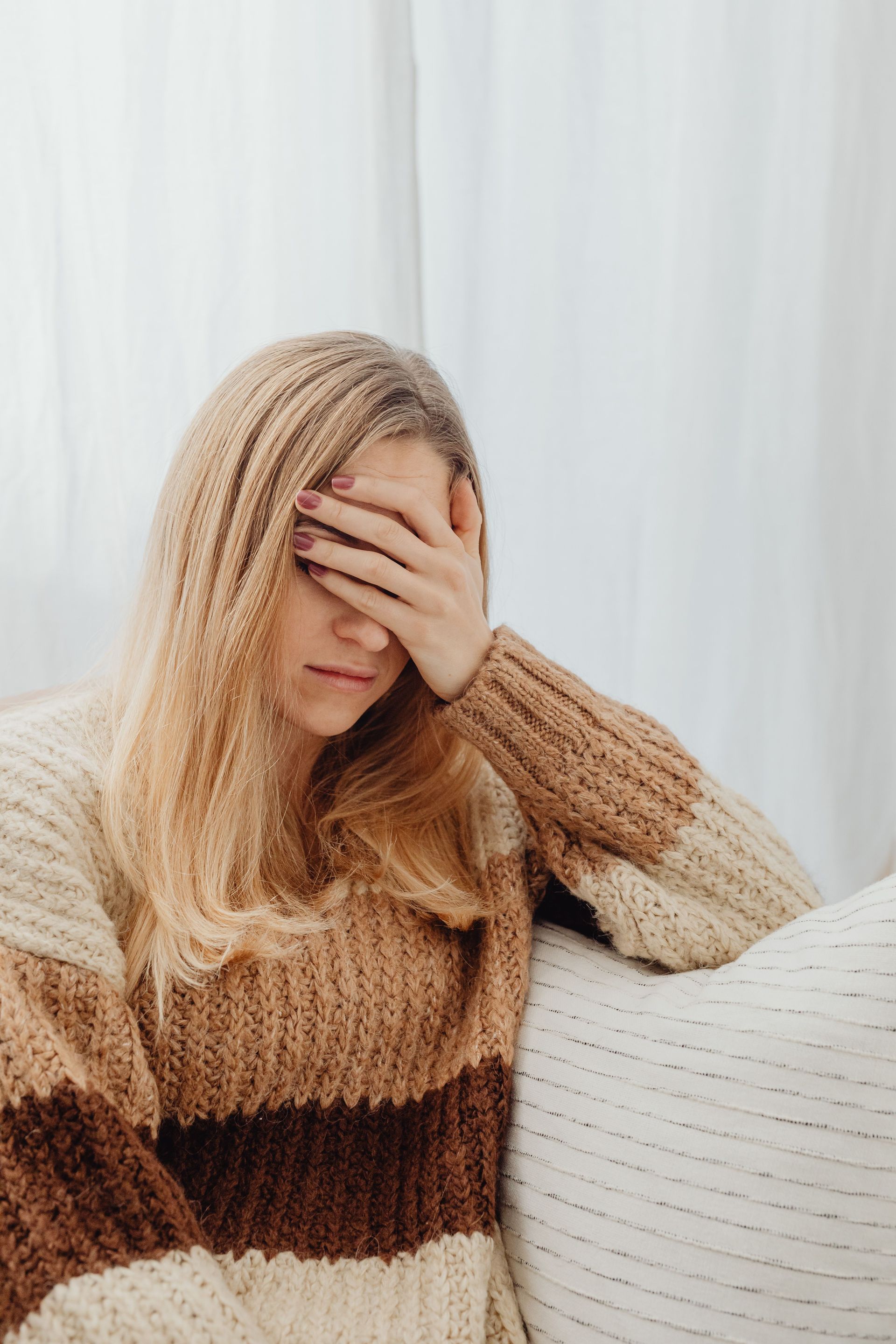 Person with hand on face, sitting indoors in a beige and brown sweater against a white curtain background