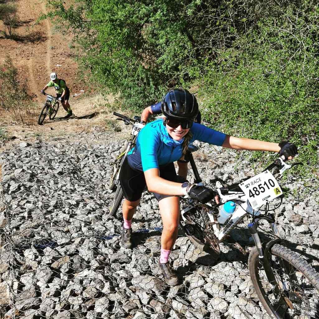 Woman pushing a mountain bike uphill on a rocky trail during a race. Other cyclists visible.