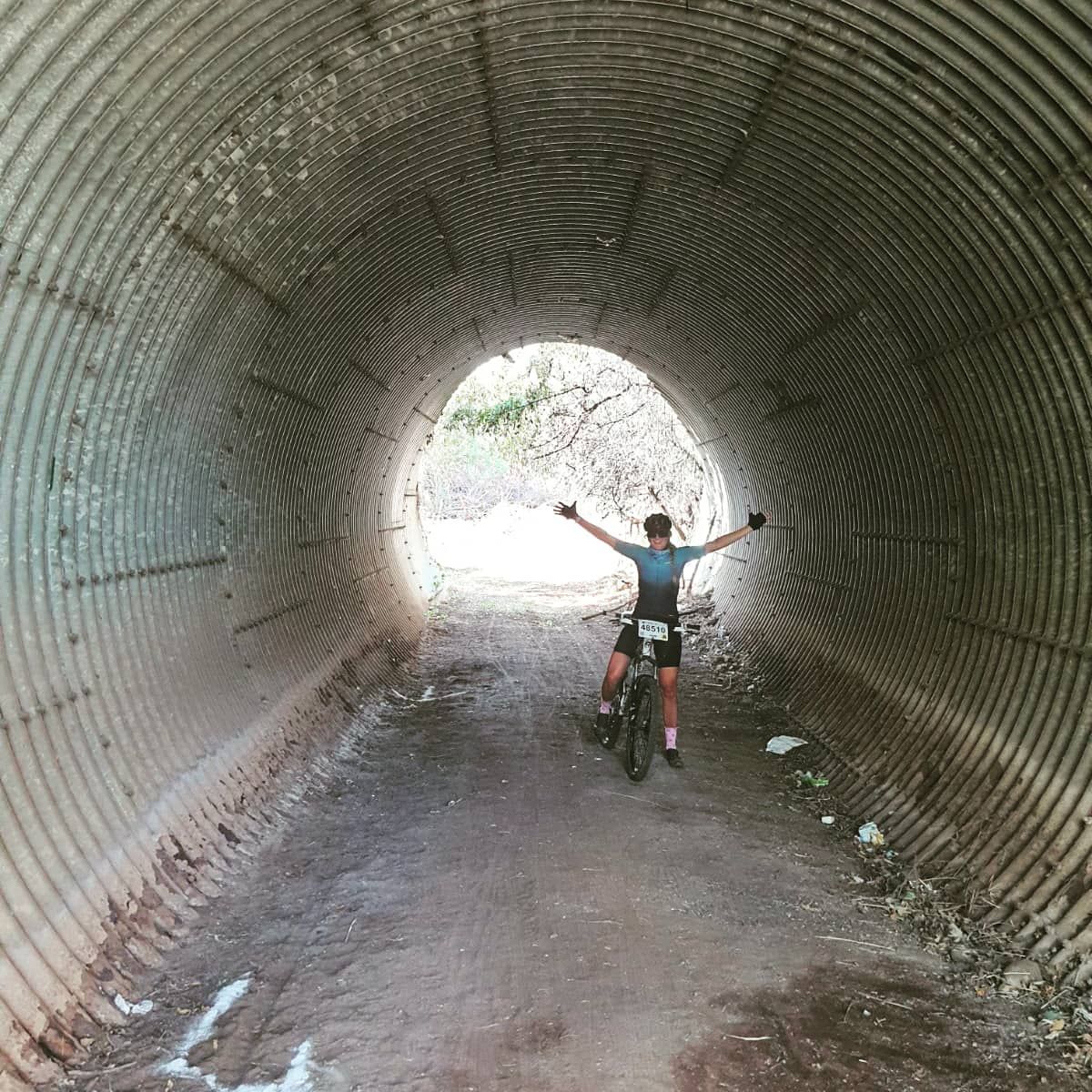 Cyclist in tunnel with arms raised, light at end. Corrugated metal walls and dirt path.