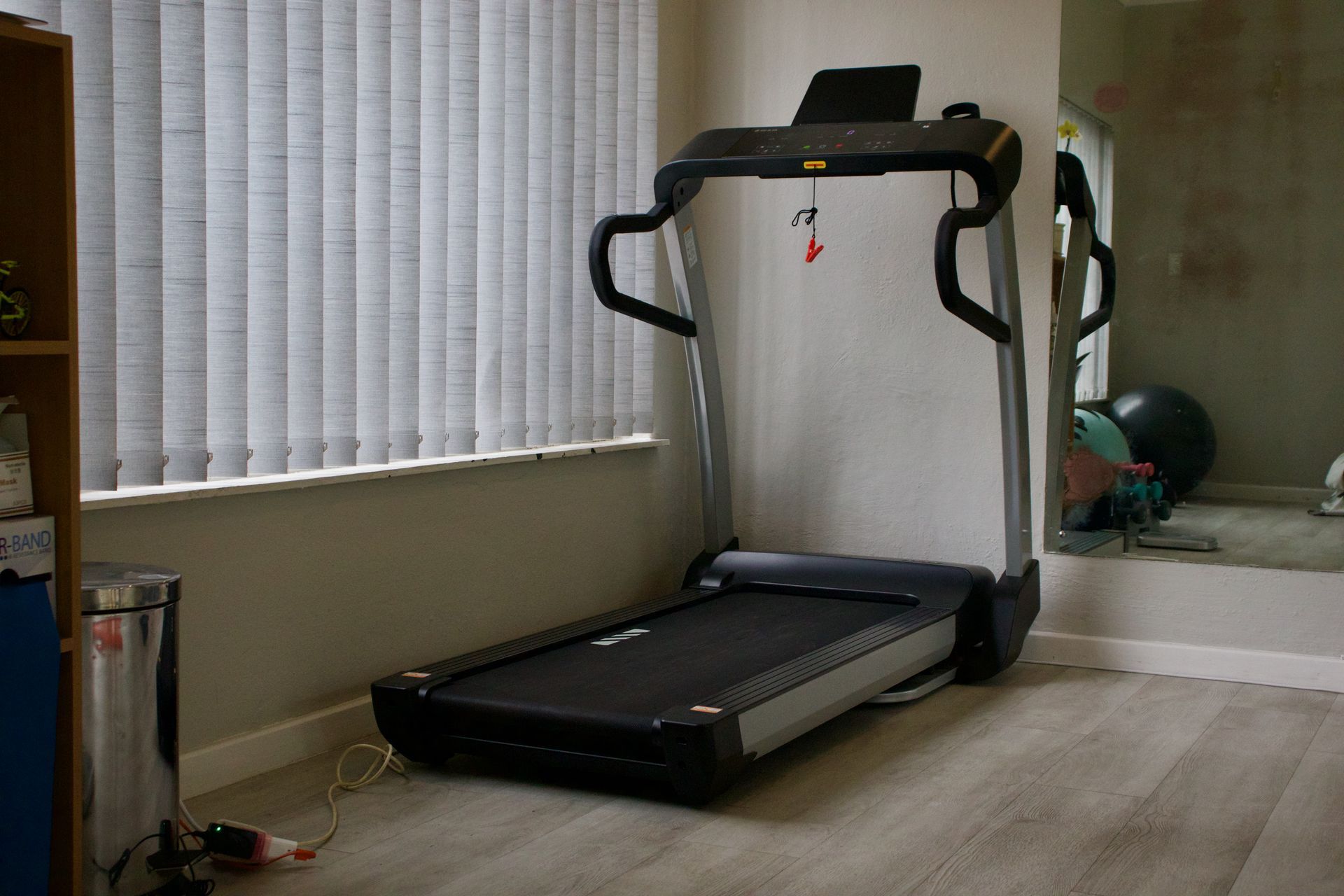 Treadmill near a window with blinds. A mirror reflects a fitness ball and other objects. Gray walls and flooring.