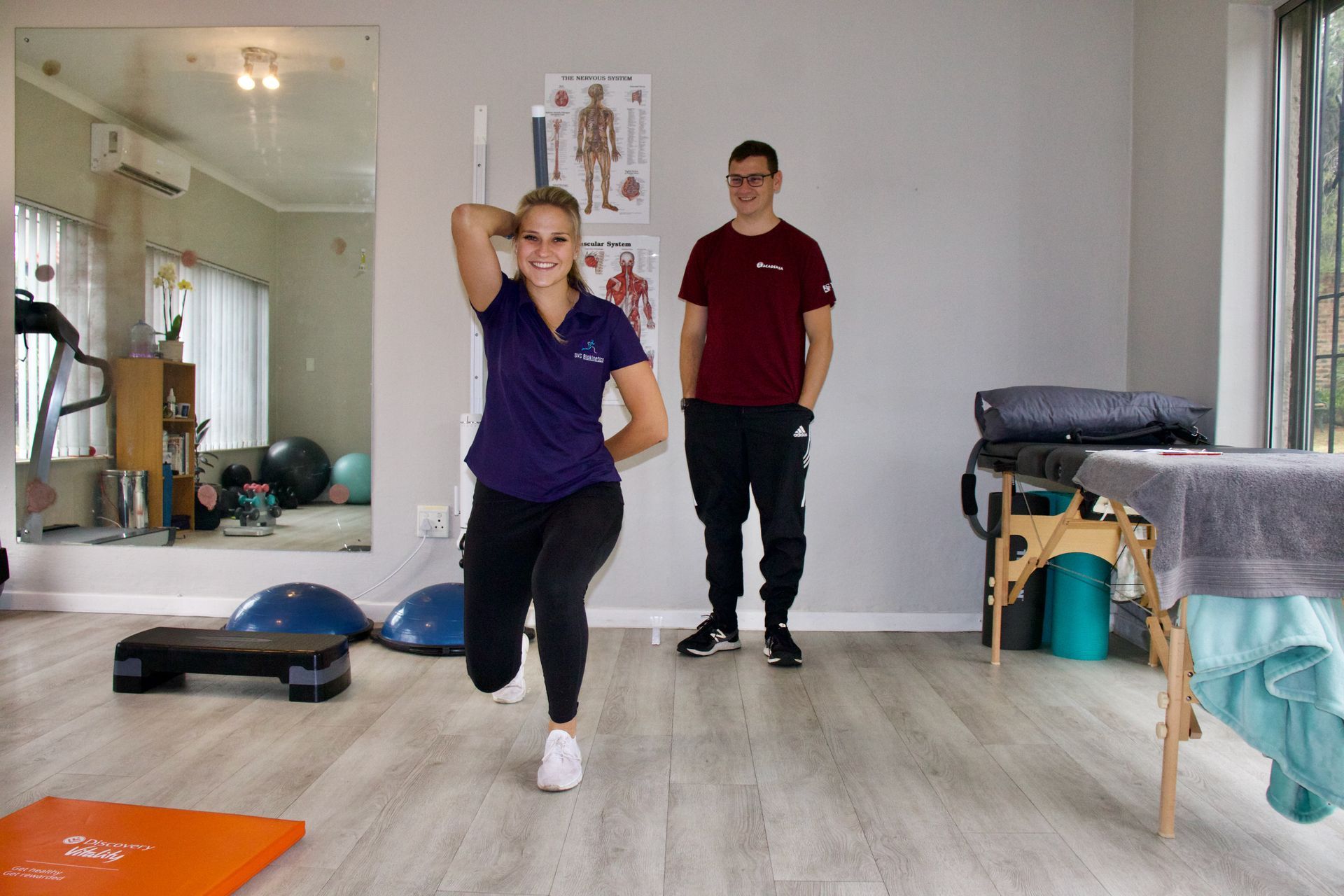 Woman doing a lunge exercise in a gym, with a man watching. White wall, exercise equipment visible.