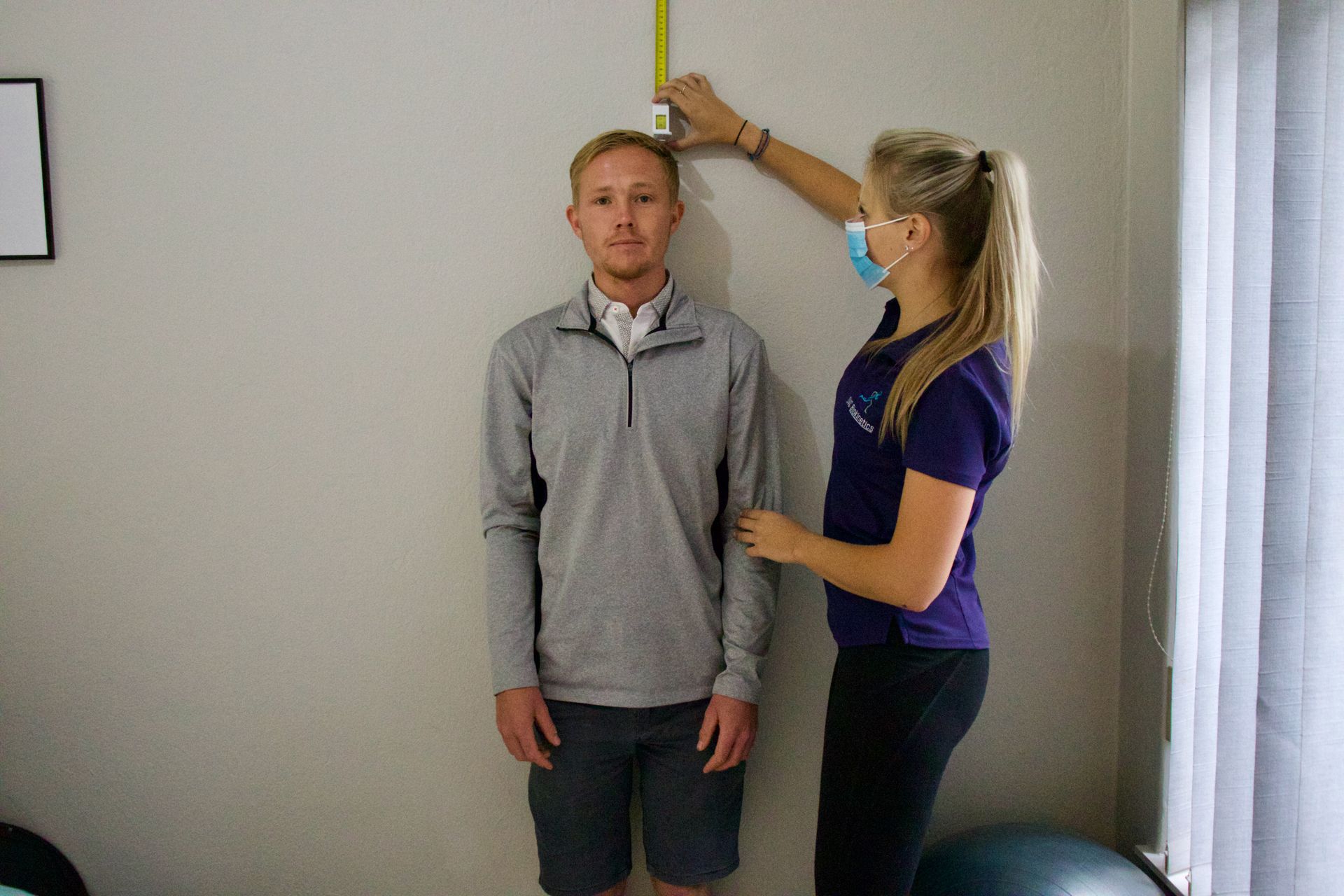Person having height measured against a wall by a person wearing a face mask.
