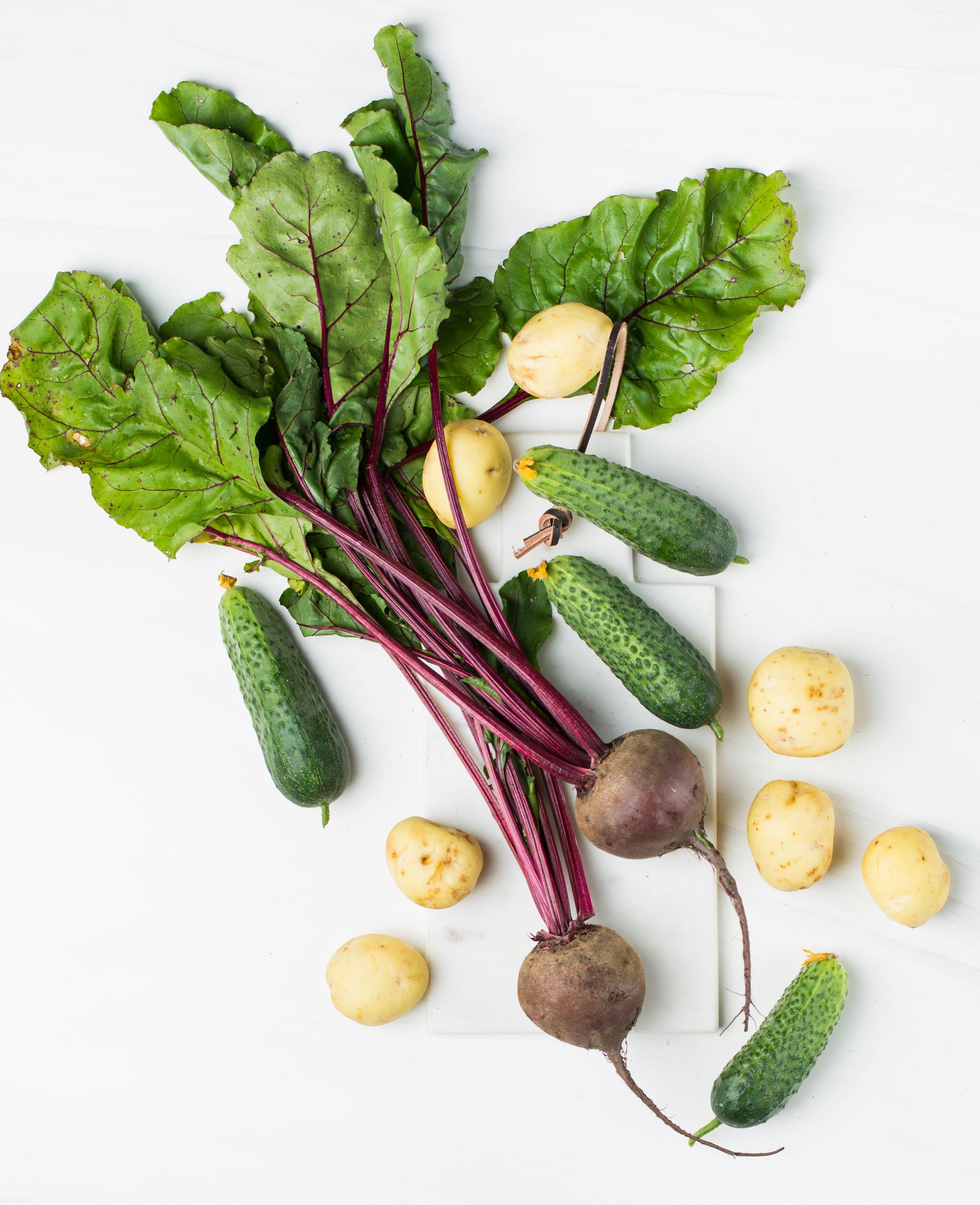 Beetroot with green leaves, potatoes, and cucumbers on a white surface.
