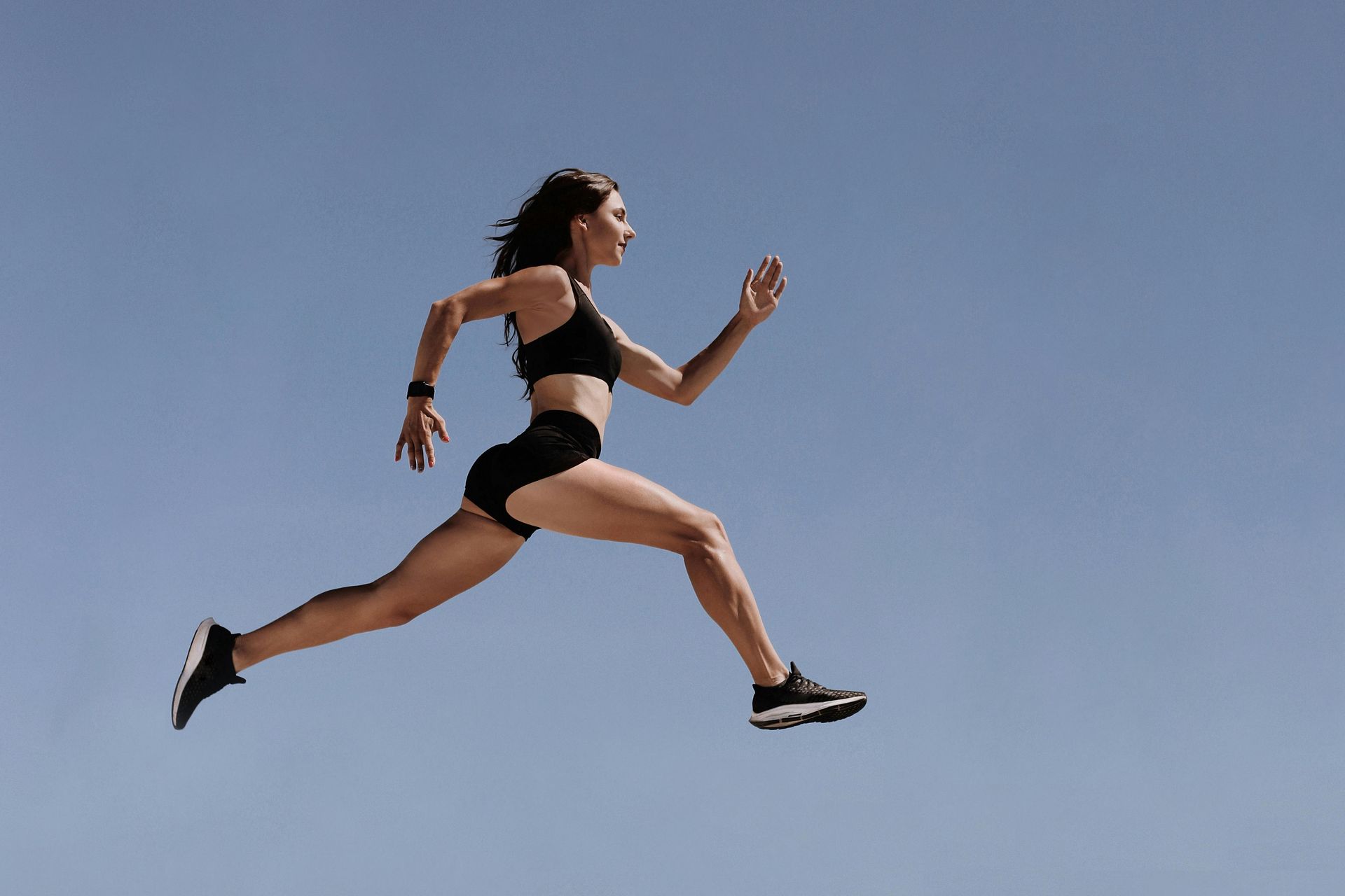 Woman in athletic wear running and jumping in mid-air against a blue sky.