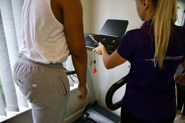 A person in sweatpants stands on a treadmill as a blonde woman in purple shirt adjusts the controls.