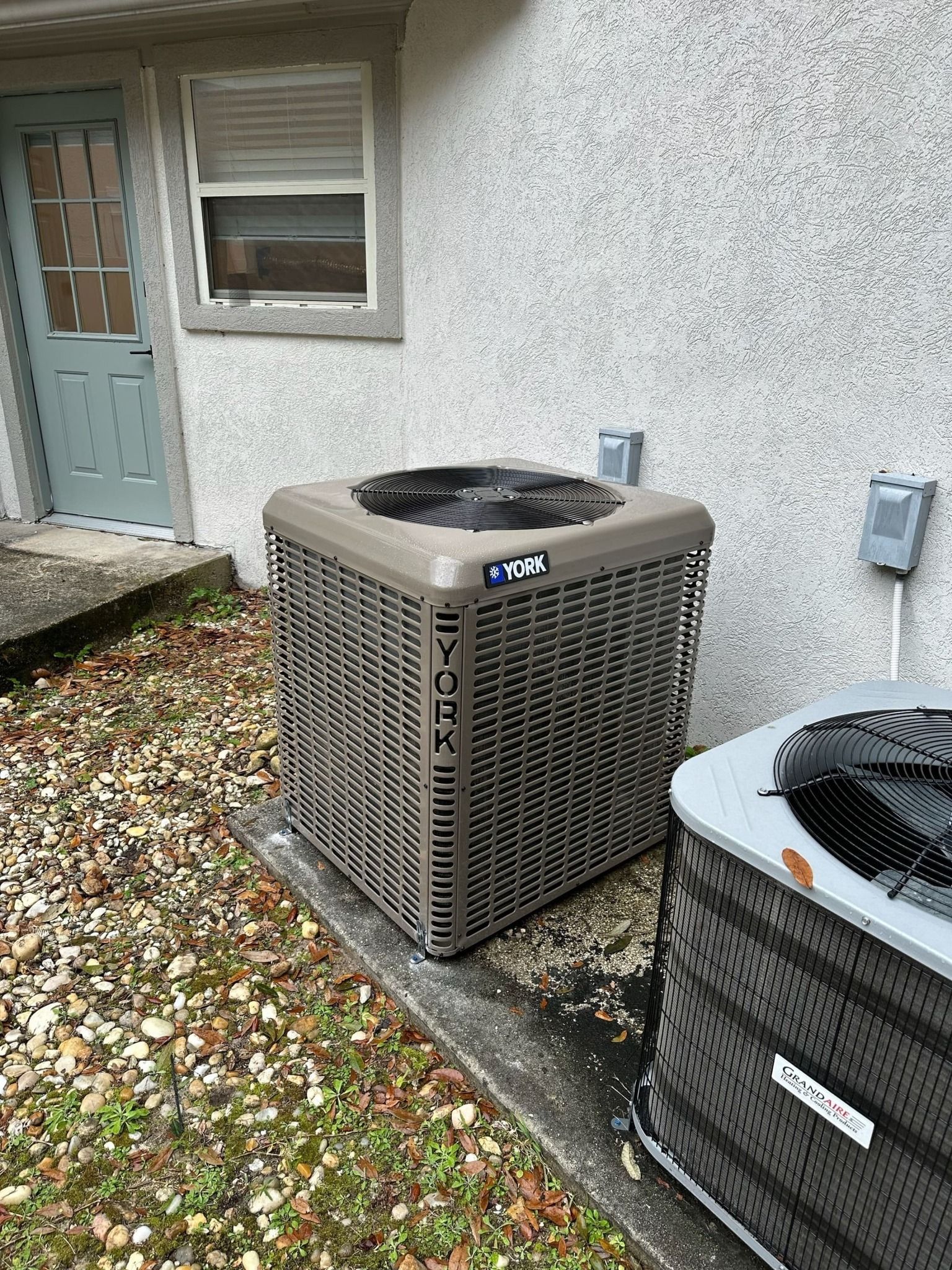 Two air conditioners are sitting next to each other on the side of a house.